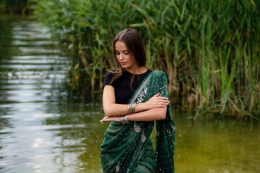 Nehir kenarında poz veren, ekstra uzun örgülü, Hint geleneksel elbiseli güzel esmer kız. Yeşil doğa arka planında, sari içinde genç ve çekici bir kadının portresi..