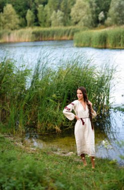 Kırsal kesimde, uzun Ukrayna elbiseli güzel esmer kız nehir kenarında kova kova su ile poz veriyor. Kırsal kesimdeki sanat fotoğraflarında oldukça genç bir model.. 