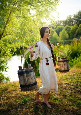 Kırsal kesimde, uzun Ukrayna elbiseli güzel esmer kız nehir kenarında kova kova su ile poz veriyor. Kırsal kesimdeki sanat fotoğraflarında oldukça genç bir model.. 
