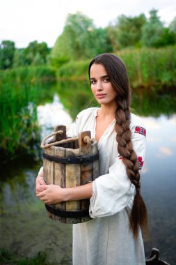 Kırsal kesimde, uzun Ukrayna elbiseli güzel esmer kız nehir kenarında kova kova su ile poz veriyor. Kırsal kesimdeki sanat fotoğraflarında oldukça genç bir model.. 