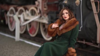 Eski moda yeşil kürklü ve kırmızı bavullu zarif siyah şapkalı, retro trenin yanında duran muhteşem kızıl saçlı bir kadının videosu. Kış istasyonundaki Leydi Yolcu. Sıcak sanat peri masalı.