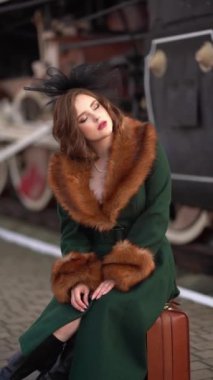 Eski moda yeşil kürklü ve kırmızı bavullu zarif siyah şapkalı, retro trenin yanında duran muhteşem kızıl saçlı bir kadının videosu. Kış istasyonundaki Leydi Yolcu. Sıcak sanat peri masalı.