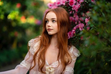 Renkli çiçeklerin yanında duran pembe klasik elbiseli ve hasır şapkalı güzel kızıl saçlı kız. Romantik bir kadının sanat eseri. Bahçede poz veren sevimli bir model..
