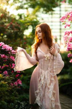 Renkli çiçeklerin yanında duran pembe klasik elbiseli ve hasır şapkalı güzel kızıl saçlı kız. Romantik bir kadının sanat eseri. Bahçede poz veren sevimli bir model..