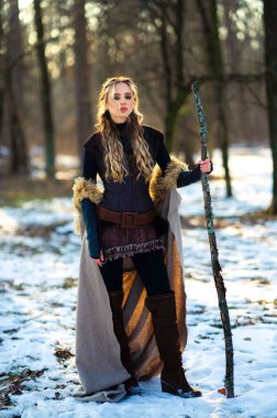 Kürklü, ortaçağ elbiseli, kış güneşli ormanlarda poz veren güzel sarışın kız. Savaşçı hakkında bir masal. Sıcak sanat eserleri.