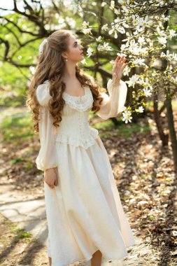 Genç sarışın kıvırcık kadın çiçek açan manolya bahçesinde yürüyor. Bahar zamanı, doğal güzellik, yumuşak güneş ışığı, romantik ve huzurlu atmosfer. Doğada güzel bir sanat portresi.