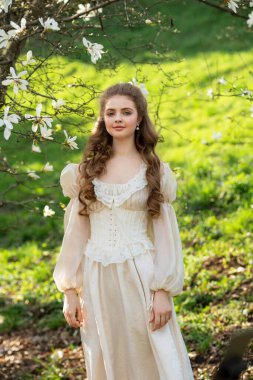 Genç sarışın kıvırcık kadın çiçek açan manolya bahçesinde yürüyor. Bahar zamanı, doğal güzellik, yumuşak güneş ışığı, romantik ve huzurlu atmosfer. Doğada güzel bir sanat portresi.