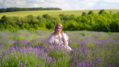 Akan mor elbiseli genç bir kadın çiçek açan çiçeklerle çevrili lavanta tarlasında zarifçe yürüyor. Yaz güneşi, romantik ruh hali ve doğal güzellik. Rüya gibi bir atmosfer ve doğada zarif bir hareket..