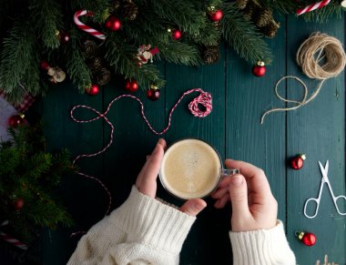 Bir Noel dekorunda, yeşil ahşap bir masanın üzerinde bir fincan kahveyi tutan kadın elleri. 