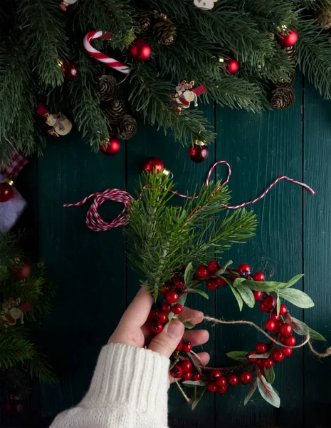 Yakın plan kadın elleri, Noel dekoru içinde yeşil ahşap bir masada Noel çelengi süslüyor.