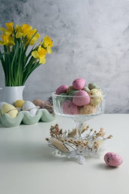 Candy sea stones in a glass near a vase with yellow daffodils and Easter eggs on the table close up