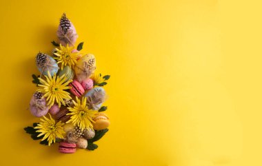 Easter eggs in a composition with yellow flowers and macaroons on a yellow background copy space