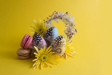 Easter eggs in a composition with yellow flowers and macaroons on a yellow background copy space