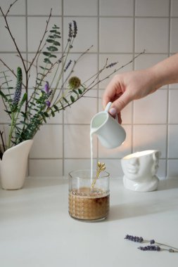 Glass texture cup with latte coffee and female hand pouring milk from jug close up