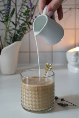 Glass texture cup with latte coffee and female hand pouring milk from jug close up