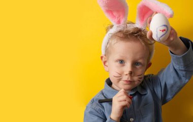 Cute boy with bunny ears shows an easter egg on a yellow background copy space. High quality photo