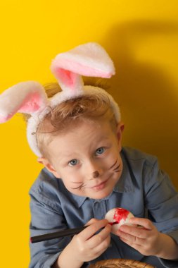 Cute boy with bunny ears paints Easter eggs on a yellow background. High quality photo