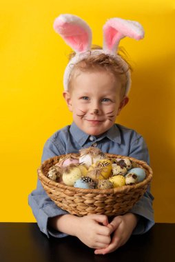 Boy with bunny ears holding basket with easter eggs on yellow background copy space. High quality 
