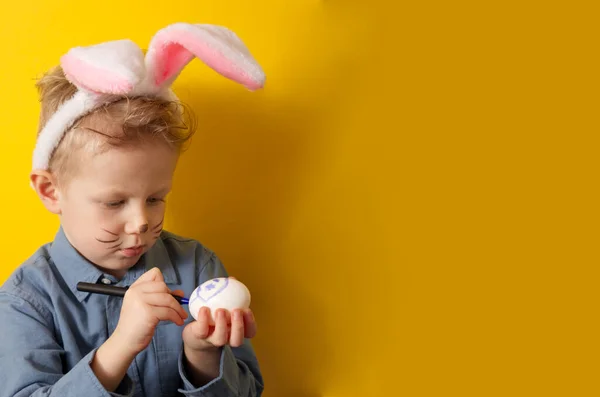 Cute boy with bunny ears paints Easter eggs on a yellow background. High quality photo