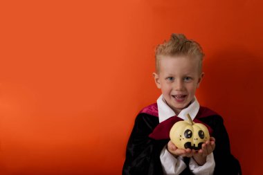 Vampir kostümlü bir çocuk turuncu arka planda balkabağı tutuyor ve gülüyor.