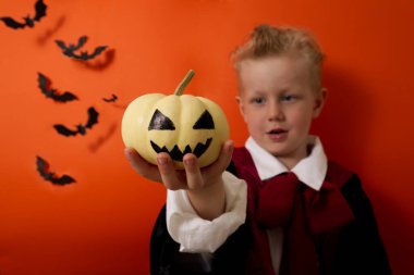 Vampir kostümlü bir çocuk turuncu arka planda balkabağı tutuyor ve gülüyor.