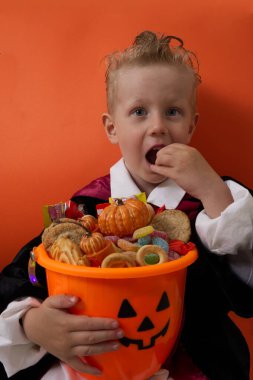 Vampir kostümlü bir çocuk turuncu arka planda bir kova dolusu şeker tutuyor.