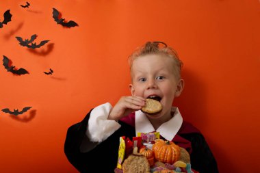 Vampir kostümlü bir çocuk turuncu arka planda bir kova dolusu şeker tutuyor.