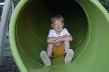 Sarışın bir çocuk oyun parkında kaydırağın içinde oturur ve gülümser.
