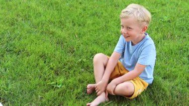 Mavi tişörtlü sarışın bir çocuk yeşil çimlerin arkasında gülüyor.