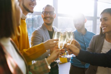 Bir grup genç iş adamı ofiste dururken birbirlerine kadeh kaldırıyor ve gülümsüyor. Ortaklar zaferlerini kutluyor. Doğum günü, yeni yıl, noel konsepti.