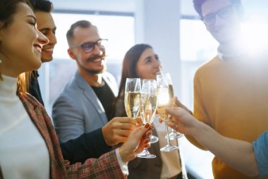 Bir grup genç iş adamı ofiste dururken birbirlerine kadeh kaldırıyor ve gülümsüyor. Ortaklar zaferlerini kutluyor. Doğum günü, yeni yıl, noel konsepti.