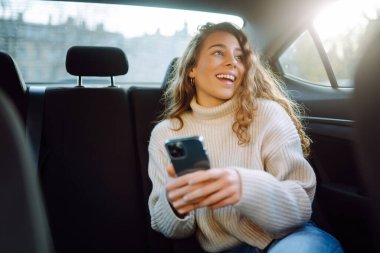 Güzel bir kadın akıllı telefon kullanıyor ve arabanın arka koltuğunda otururken gülümsüyor. İş, teknoloji, blog kavramı.
