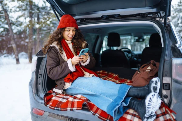 年轻女人坐在汽车行李箱上 通过手机传递信息 在阳光充足的冬季森林里舒服地交流生意 生活方式概念 — 图库照片