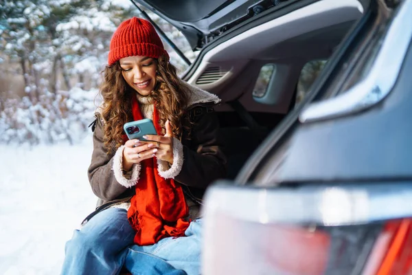 年轻女人坐在汽车行李箱上 通过手机传递信息 在阳光充足的冬季森林里舒服地交流生意 生活方式概念 — 图库照片