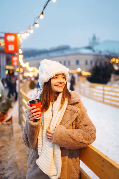 stock image Happy woman with disposable paper coffee cup in winter over outdoor ice skating rink on background. Christmas, hot drinks and holidays concept.