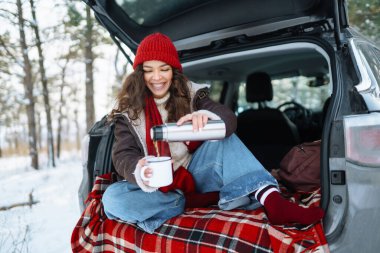 Mutlu kadın bir termos tutuyor ve kış ormanlarında arabanın bagajında çay içiyor. Dinlenme, rahatlama, seyahat, yaşam tarzı konsepti.