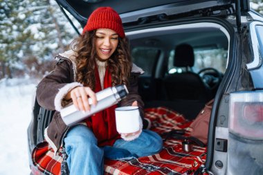 Mutlu kadın bir termos tutuyor ve kış ormanlarında arabanın bagajında çay içiyor. Dinlenme, rahatlama, seyahat, yaşam tarzı konsepti.