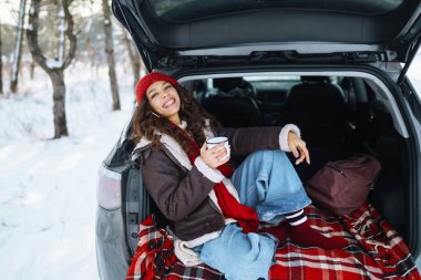 Mutlu kadın bir termos tutuyor ve kış ormanlarında arabanın bagajında çay içiyor. Dinlenme, rahatlama, seyahat, yaşam tarzı konsepti.