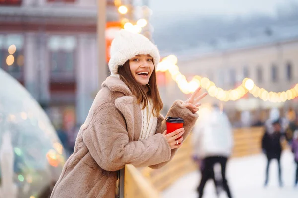 Young woman with coffee in winter over outdoor ice skating rink on background. Festive Christmas fair, winter holidays concept.
