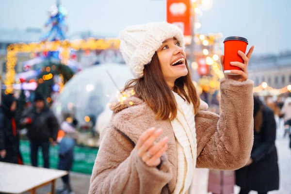 Young woman with coffee in winter over outdoor ice skating rink on background. Festive Christmas fair, winter holidays concept.