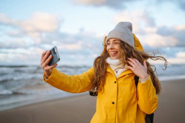 Telefonu olan genç bir turist. Selfie zamanı. Güneş batarken deniz kenarında poz veren sarı ceketli bir turist. Seyahat, yaşam tarzı, macera.