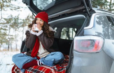 Genç kadın bir termos tutuyor ve kış ormanlarında arabanın bagajında çay içiyor..