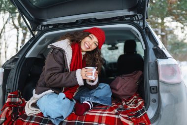 Genç kadın bir termos tutuyor ve kış ormanlarında arabanın bagajında çay içiyor..