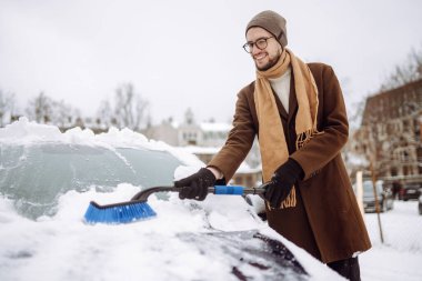 Kış kar yağışı sırasında arabasındaki karları temizleyen gülümseyen adamın portresi. Ulaşım, kış, hava, insan ve araç kavramı.