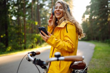 Pretty young womanusing mobile phone in the city while listening music through earphones to go by the bicycle in the park. Lifestyle. Relax, nature concept.