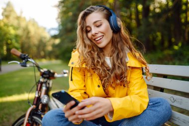 Pretty young womanusing mobile phone in the city while listening music through earphones to go by the bicycle in the park. Lifestyle. Relax, nature concept.
