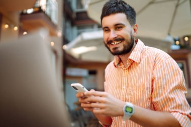 Bilgisayarı ve telefonu olan genç bir adam bir kafede oturuyor ve çalışıyor. İş, blog yazma, serbest çalışma, eğitim kavramı.