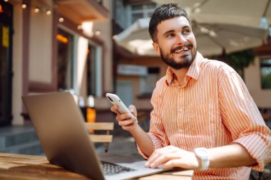 Bilgisayarı ve telefonu olan genç bir adam bir kafede oturuyor ve çalışıyor. İş, blog yazma, serbest çalışma, eğitim kavramı.