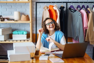 Genç bayan tasarımcı iş yerinde dizüstü bilgisayarla oturuyor. Küçük işletme sahibi, girişimci, ecommerce giyim mağazasının siparişlerini kontrol eden satıcı. Alışveriş konsepti. Moda stüdyosu.
