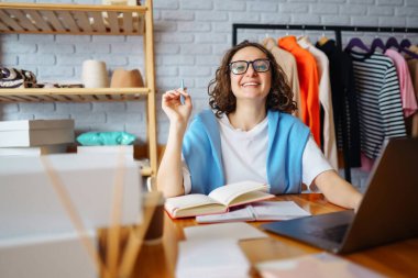 Genç bayan tasarımcı iş yerinde dizüstü bilgisayarla oturuyor. Küçük işletme sahibi, girişimci, ecommerce giyim mağazasının siparişlerini kontrol eden satıcı. Alışveriş konsepti. Moda stüdyosu.